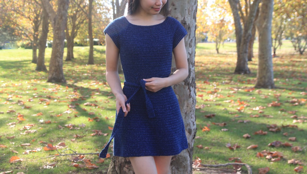 woman standing in front of a tree wearing a blue crochet dress with belt