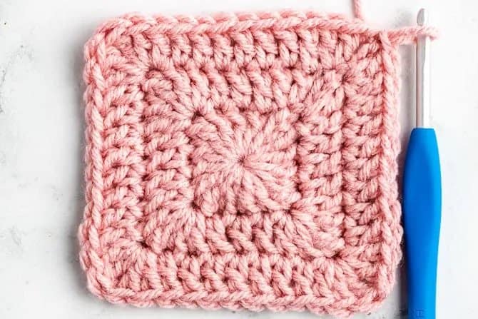 Solid Granny Square in pink beside a crochet needle